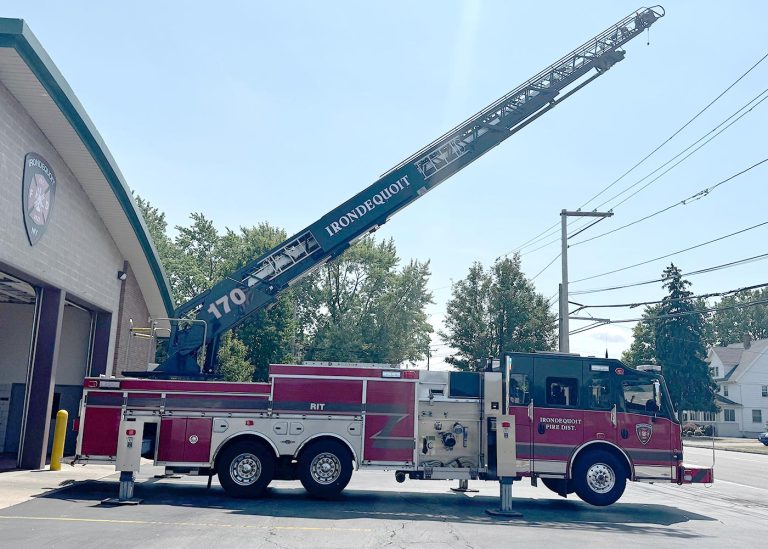 A red and white fire truck labeled "IRONDQUOIT" with an extended ladder is parked outside a fire station on a sunny day. Trees and utility poles are visible in the background.