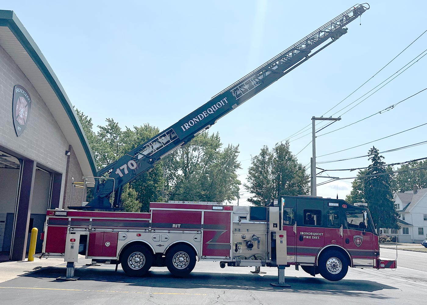 A red and white fire truck labeled "IRONDQUOIT" with an extended ladder is parked outside a fire station on a sunny day. Trees and utility poles are visible in the background.