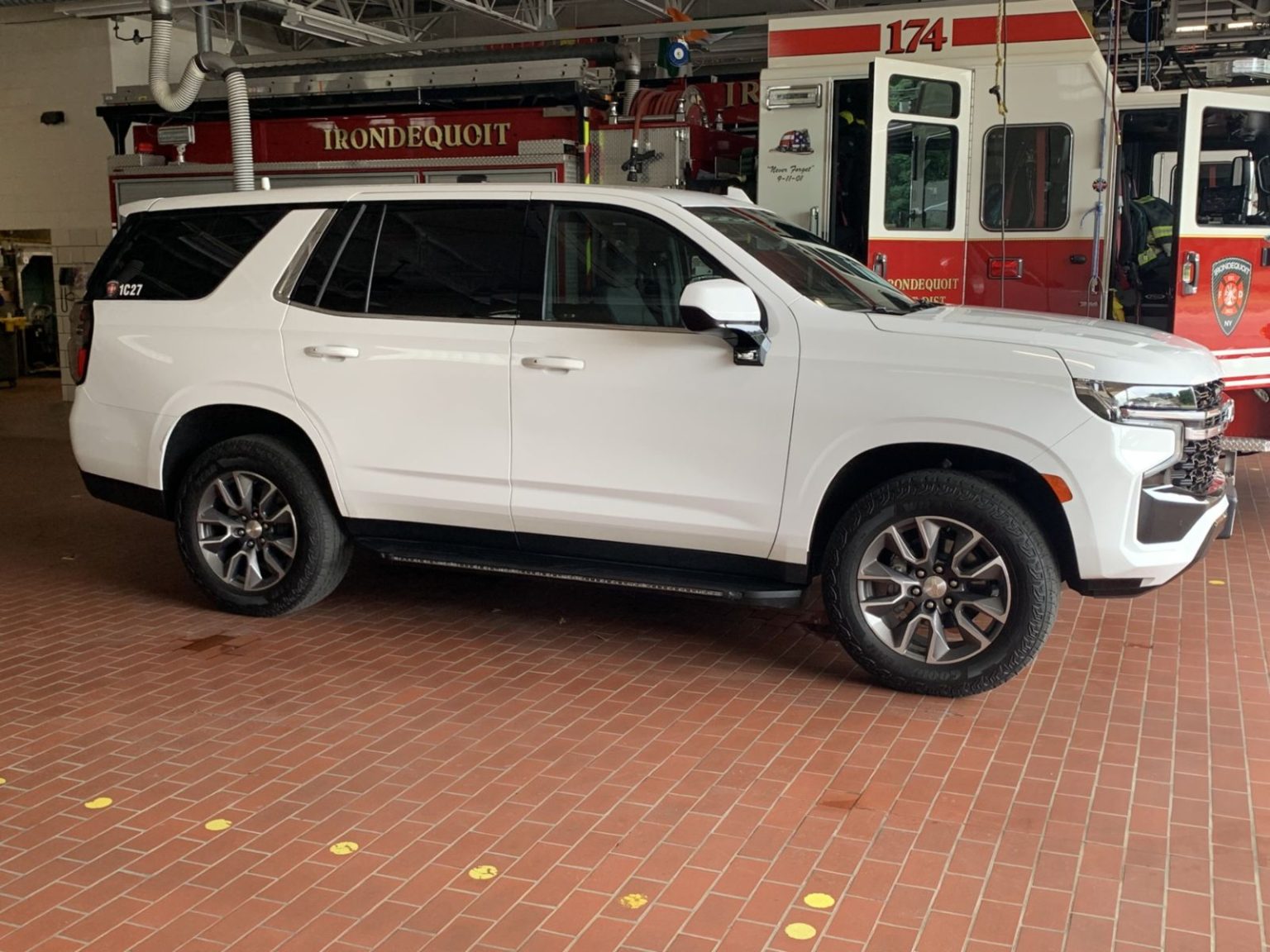A white SUV parked indoors on a brick floor, with fire trucks and emergency vehicles labeled "IRONDEQUOIT" and "174" visible in the background.