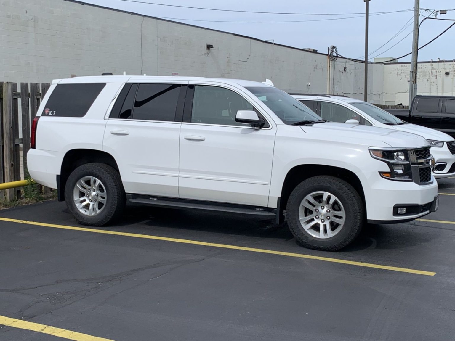 A white Chevrolet Tahoe SUV is parked in a blacktop parking lot next to other vehicles, with a fence and industrial buildings in the background.