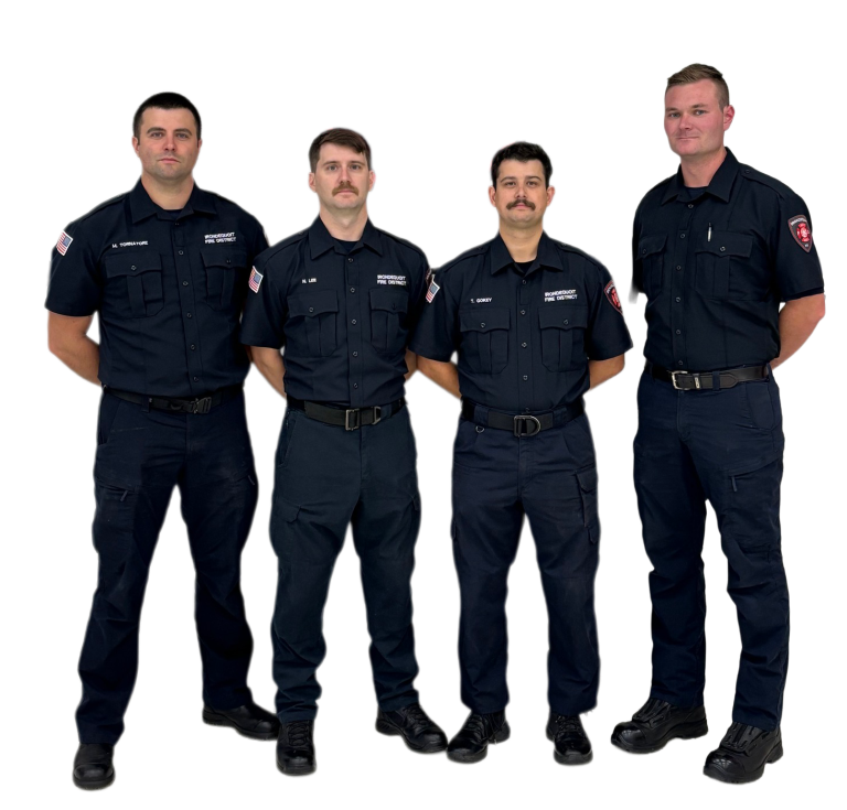 Four firefighters stand in a row, wearing matching dark blue uniforms with badges and name tags, hands behind their backs, against a plain white background.