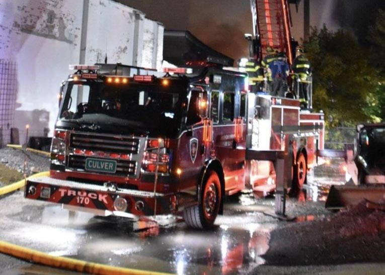 A fire truck labeled "Culver" is parked near a building at night, with firefighters climbing a ladder as smoke rises in the background. Reflective surfaces show emergency lights and wet ground.