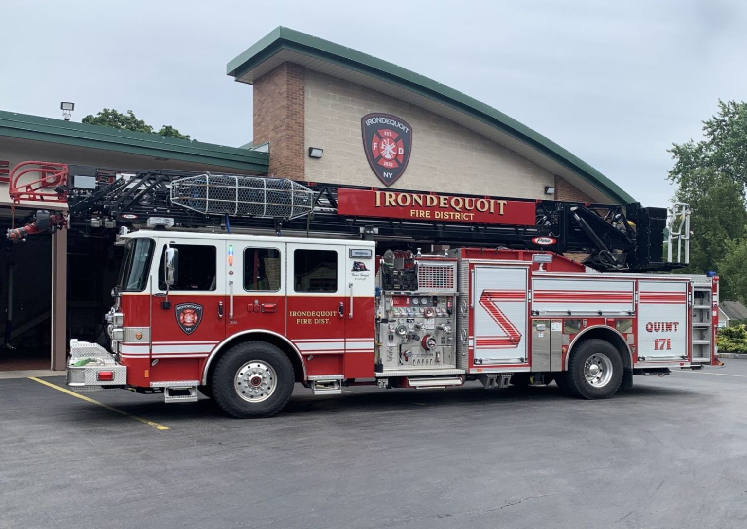A red and white Ironddequoit Fire District fire truck with a large ladder is parked outside a fire station building. The truck is labeled "QUINT 171.