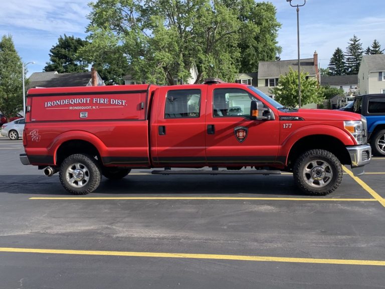 A red Ironddequoit Fire District truck is parked in a lot with yellow lines. The truck has emergency lights on top and is marked with department insignia. Trees and houses are visible in the background.