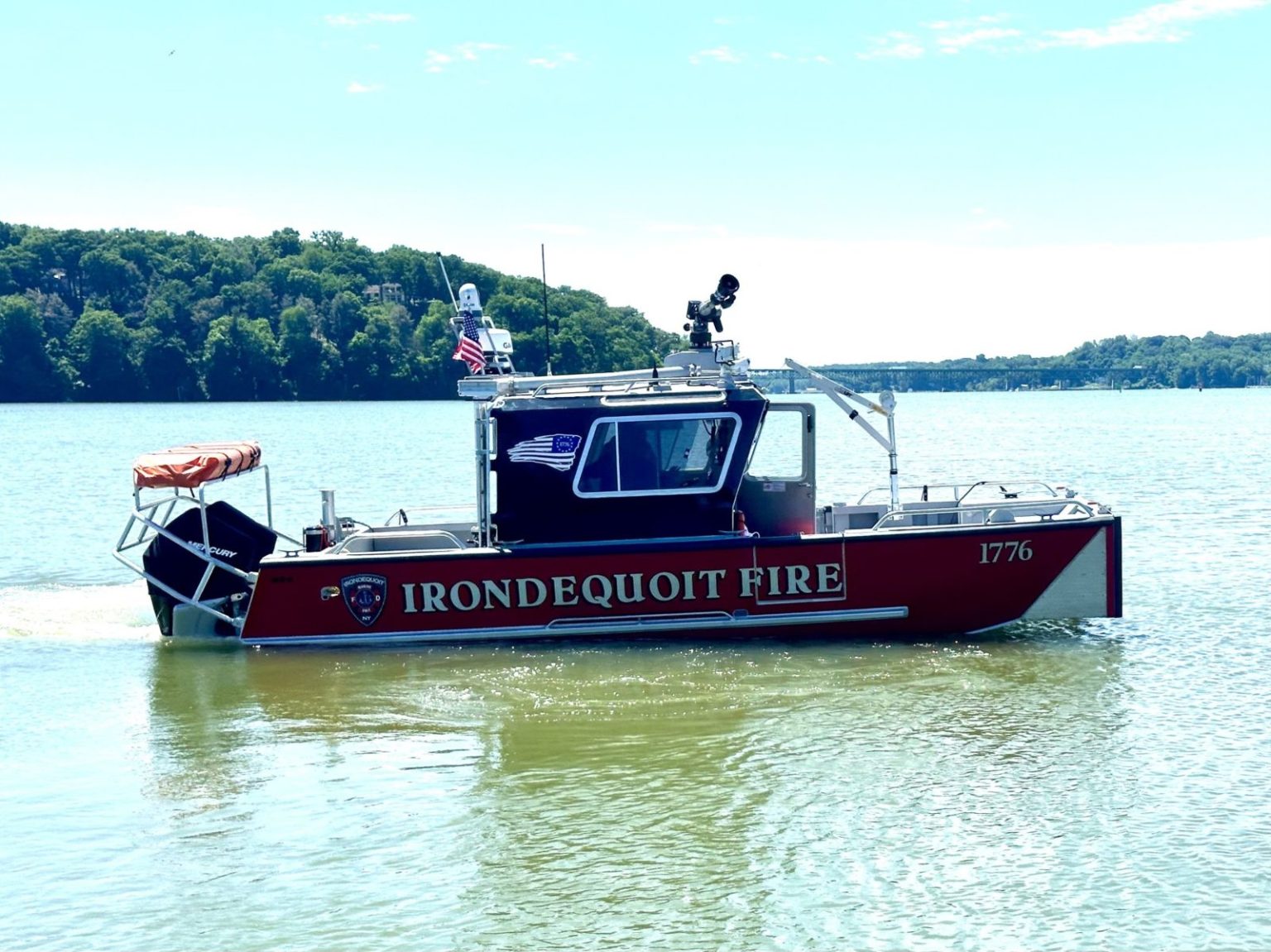 A red and silver fire rescue boat labeled "IRONDEQUOIT FIRE" sails on a calm lake, with trees in the background and a clear sky above. The boat has emergency equipment and an American flag on top.