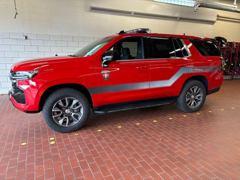A red fire department SUV with gray stripes is parked indoors on a brick floor next to fire equipment and a white tiled wall.