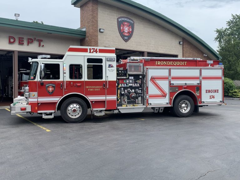 A red and white fire truck labeled “IRONDQUOIT” and “ENGINE 174” is parked outside a fire station garage with “DEPT.” on the building. The fire truck has various equipment compartments and emergency lights.