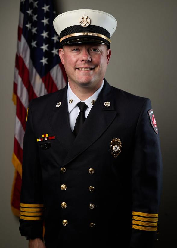 A smiling firefighter in formal dress uniform and white hat stands in front of an American flag. The uniform includes badges, medals, and a shoulder patch, indicating a high rank.