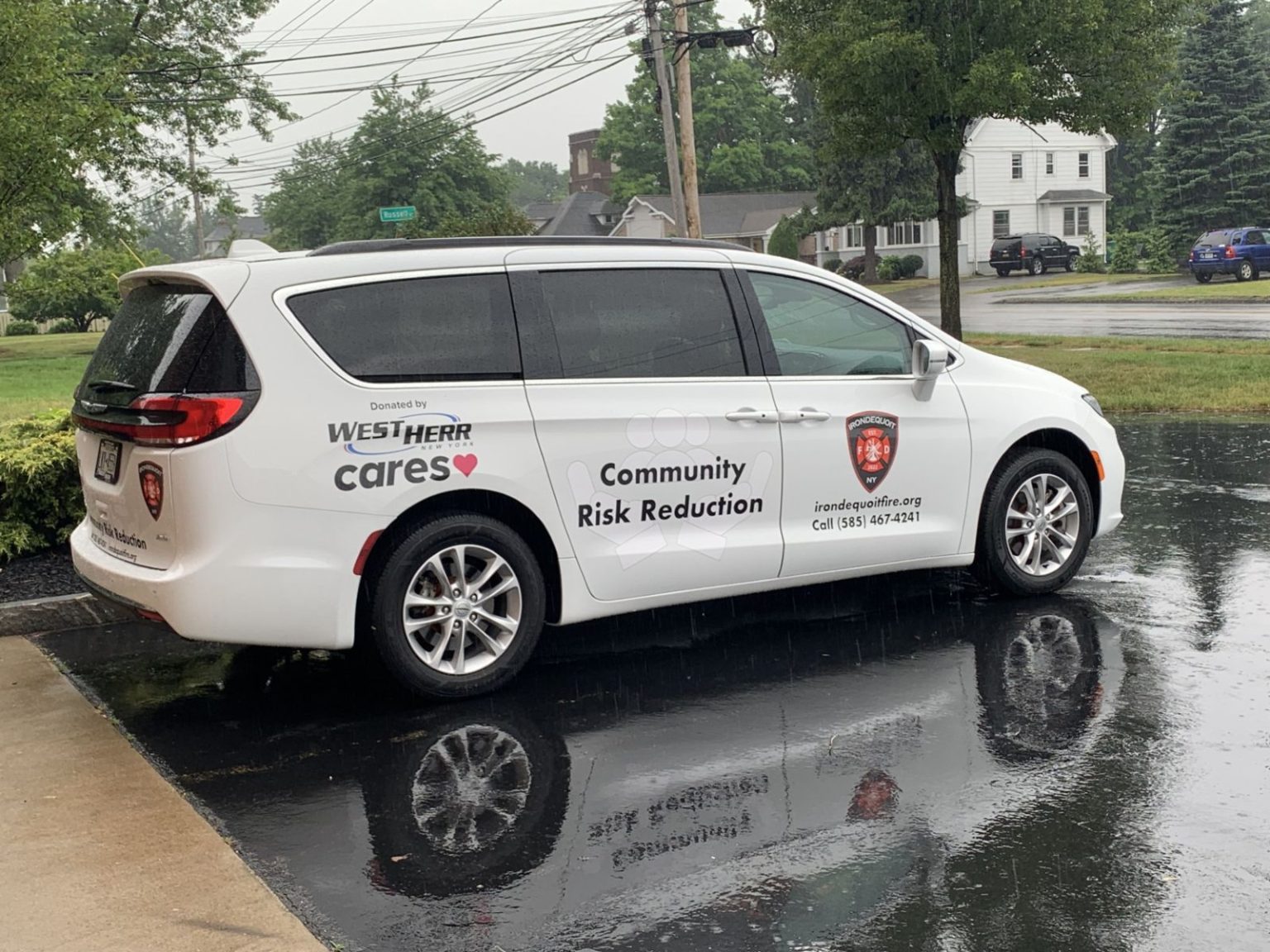 A white minivan parked on wet pavement, displaying "Community Risk Reduction" and a fire department logo on its side, with text indicating it was donated by West Herr Cares. Trees and utility poles are in the background.