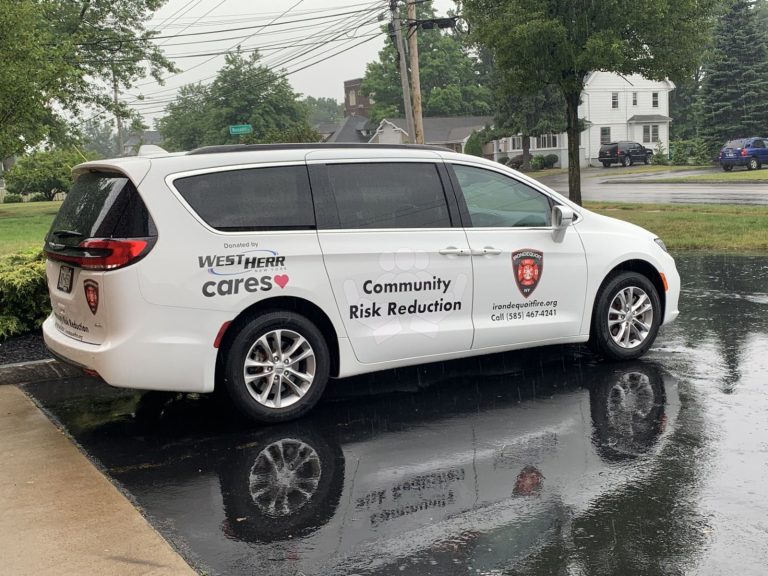 A white minivan parked on wet pavement, displaying "Community Risk Reduction" and a fire department logo on its side, with text indicating it was donated by West Herr Cares. Trees and utility poles are in the background.