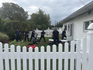 Seven emergency responders in navy uniforms work together using ropes and equipment beside a white house and white picket fence, with a stretcher and medical bags on the lawn under a cloudy sky.