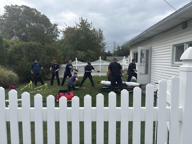 Seven emergency responders in navy uniforms work together using ropes and equipment beside a white house and white picket fence, with a stretcher and medical bags on the lawn under a cloudy sky.