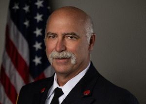 A man in a formal dark fire department uniform with badges stands in front of a U.S. flag, smiling with a mustache, against a plain background.