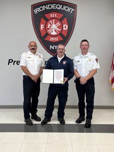 Three firefighters stand indoors in front of an Irondequoit Fire Department emblem. The middle firefighter holds a framed certificate; all are dressed in uniform and smiling. An American flag is visible on the right.