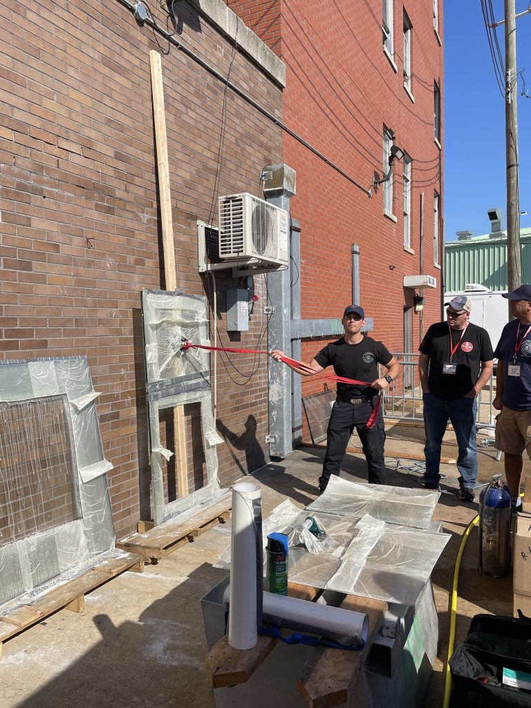 A person in black clothing uses a red sledgehammer to strike a glass window set in a brick wall, while two others watch nearby in an alleyway with construction materials and equipment scattered around.