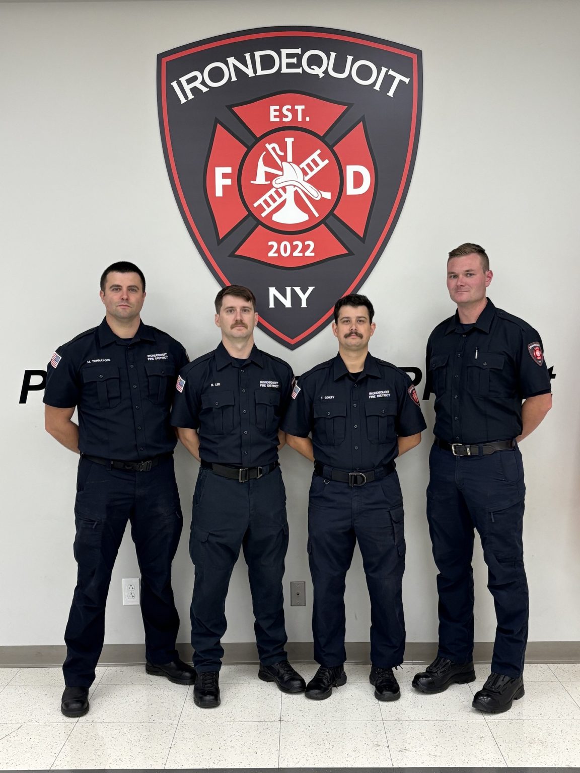 Four firefighters in uniform stand in front of a large Irondequoit Fire Department emblem on a wall, posing for a group photo.