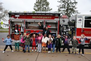A group of young children and an adult stand in front of an open fire truck, smiling and making playful faces, with trees and houses in the background on a cloudy day.