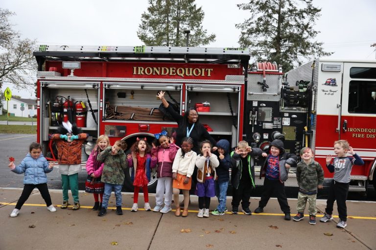 A group of young children and an adult stand in front of an open fire truck, smiling and making playful faces, with trees and houses in the background on a cloudy day.