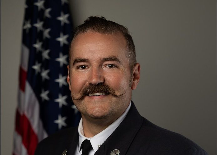 A firefighter in formal uniform with a badge, mustache, and neatly styled hair stands smiling in front of an American flag and a gray background. The patch on his sleeve reads "Irondequoit Fire" and "NY.