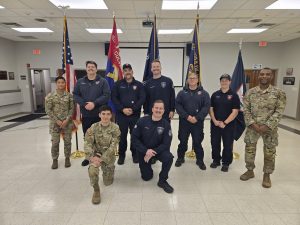Nine people in military and firefighter uniforms pose for a group photo indoors. Behind them are several flags, including the U.S. flag, in a well-lit room with tiled floors and a white ceiling.