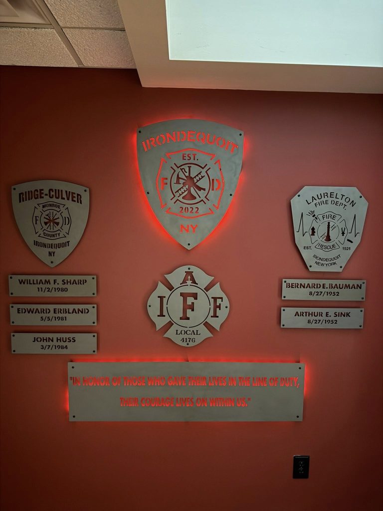 Five engraved plaques on a red wall honor local firefighters, with names, departments, and dates. A central illuminated Irondequoit Fire Department emblem and a quote plaque pay tribute to fallen heroes.