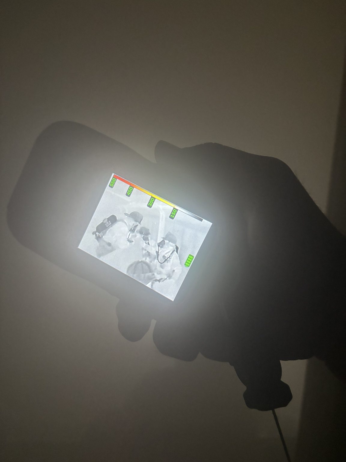 A hand holds a thermal imaging device displaying a black-and-white heat signature image of people, with colored indicators at the top of the screen, against a dark background.
