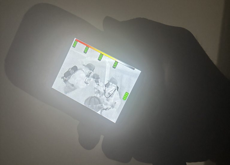 A hand holds a thermal imaging device displaying a black-and-white heat signature image of people, with colored indicators at the top of the screen, against a dark background.