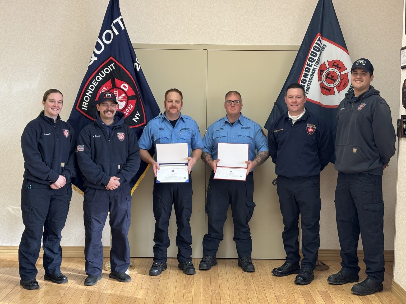 Six firefighters pose indoors, two holding framed certificates. They stand in front of two Irondequoit Fire District flags, smiling and wearing navy blue uniforms and badges. The setting is well-lit with a wooden floor.
