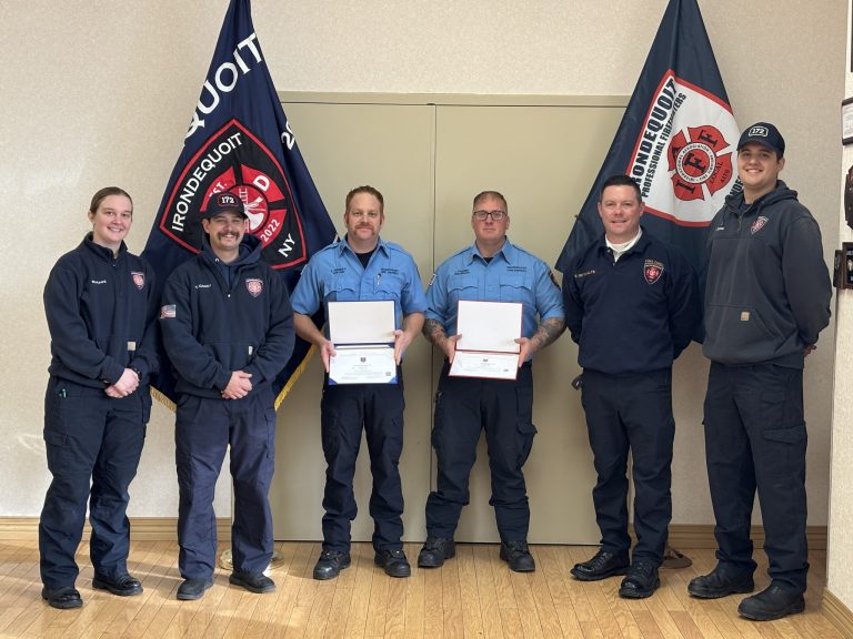 Six firefighters pose indoors, two holding framed certificates. They stand in front of two Irondequoit Fire District flags, smiling and wearing navy blue uniforms and badges. The setting is well-lit with a wooden floor.