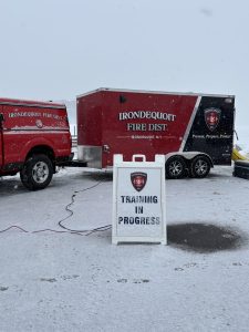 A snowy scene with a red fire district truck and trailer. In the foreground, a sign reads “TRAINING IN PROGRESS” with the Irondequoit Fire District logo. Snow covers the ground and vehicles.