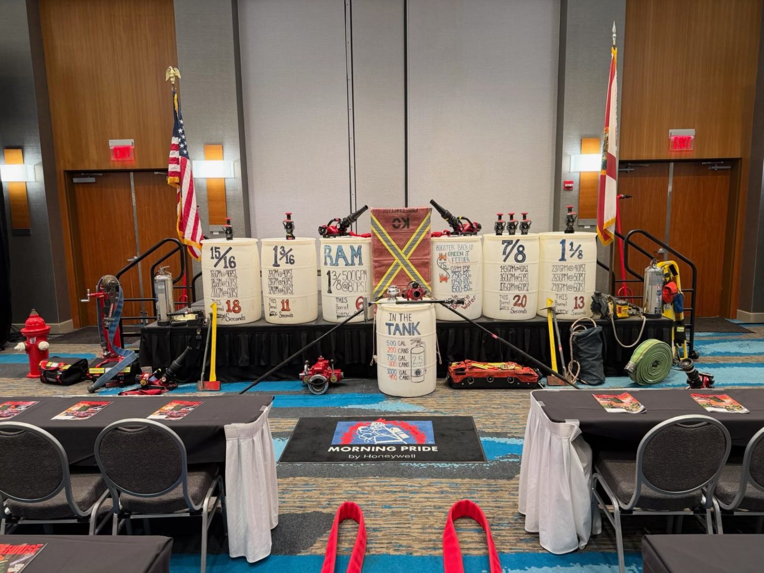 A conference room stage displays firefighting equipment, water barrels with markings, hoses, hydrants, and U.S. and state flags. Tables and chairs face the stage, and a "Morning Pride" logo is on the carpet in front.