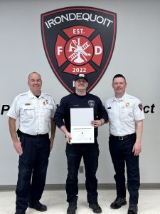 Three uniformed firefighters stand in front of an Irondequoit Fire Department emblem. The firefighter in the center holds a certificate, flanked by two others smiling at the camera.