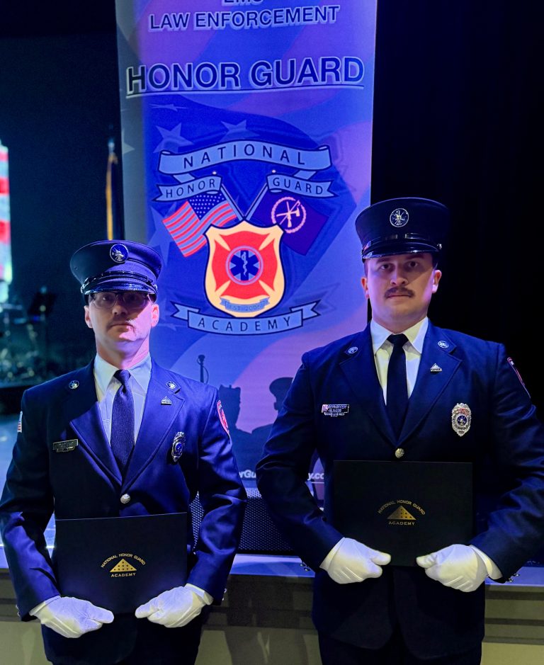 Two uniformed officers stand side by side holding certificates in front of a “National Honor Guard Academy” banner at a law enforcement event. Both wear dress hats, ties, and white gloves.